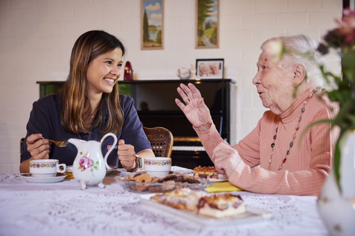 Junge Ehrenamtlerin des Besuchsdienstes sitzt mit einer Seniorin bei Kaffee und Kuchen zusammen.